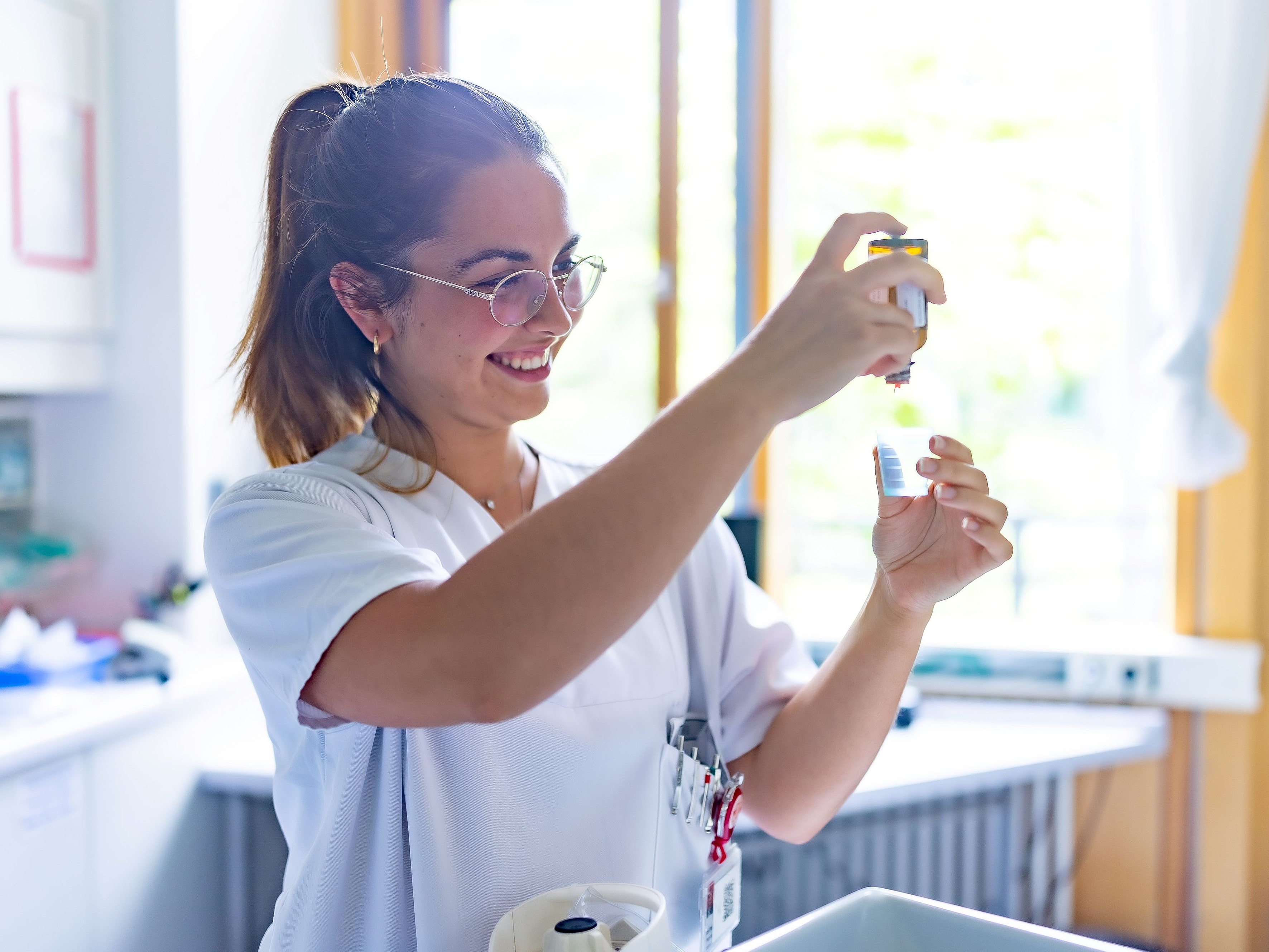 Konzentrierte Pflegekraft bei der Medikamentenvorbereitung Eine lächelnde junge Frau in einem weißen Kasack mit Brille hält konzentriert zwei kleine Behälter in den Händen, zur Vorbereitung von Medikamenten.Eine lächelnde junge Frau in einem weißen Kasack mit Brille hält konzentriert zwei kleine Behälter in den Händen, zur Vorbereitung von Medikamenten.